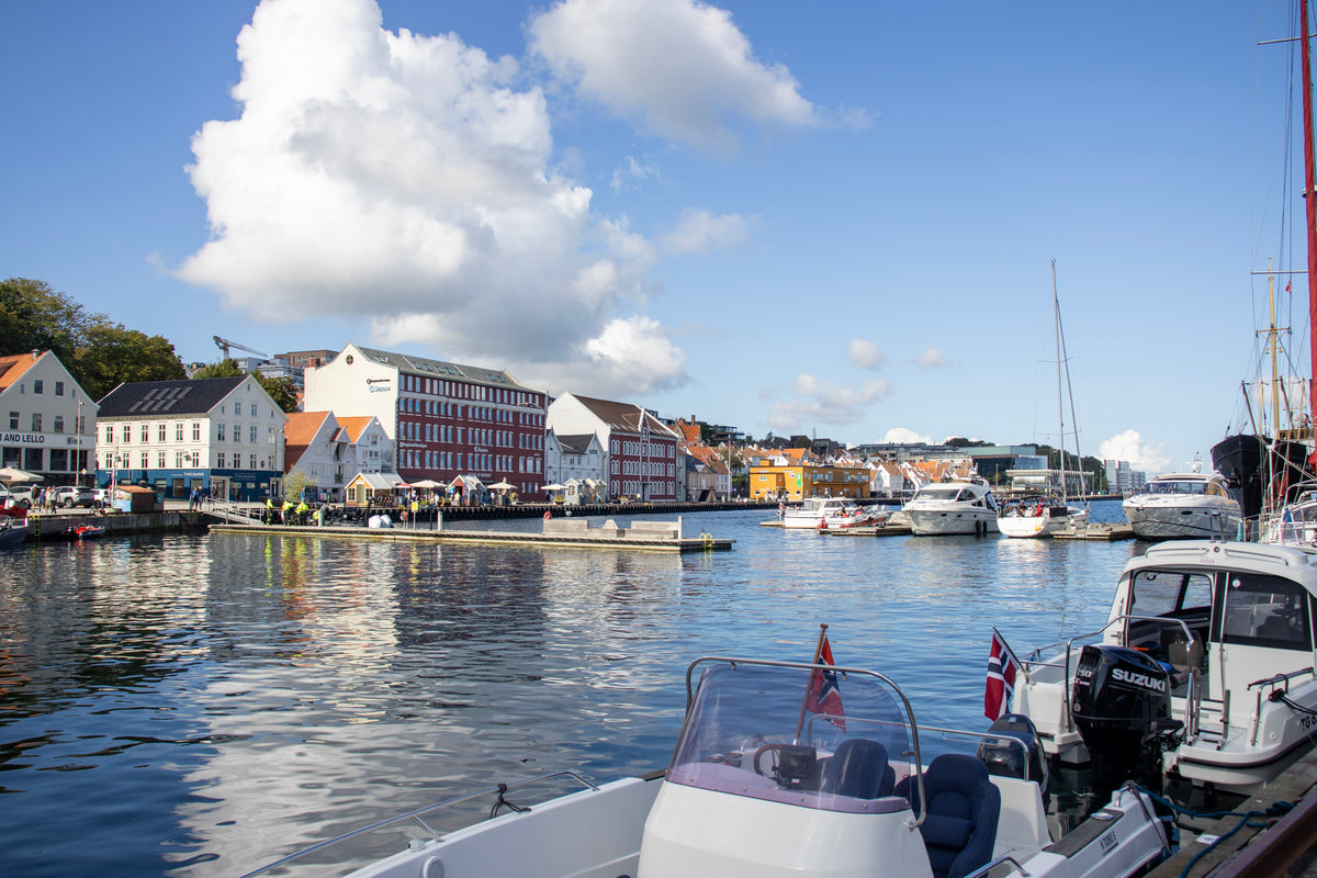 A scenic harbor view with colorful waterfront buildings, blue water, and several boats docked in the foreground under a bright blue sky with large white clouds.